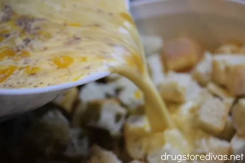 A yellow liquid being poured onto bread cubes in a white casserole pan.