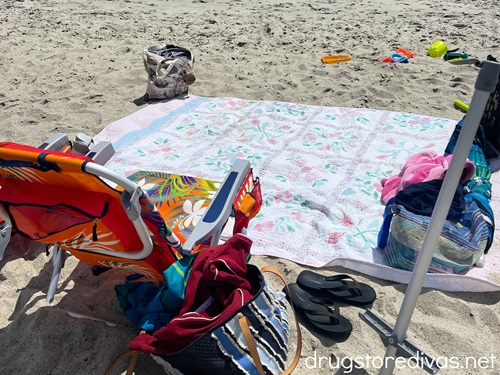 A beach blanket on the beach surrounded by beach chairs and bags.
