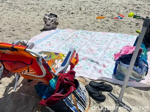 A beach blanket on the beach surrounded by beach chairs and bags.