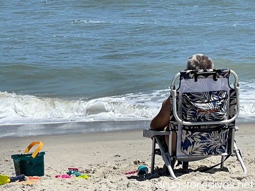 A man sitting in a beach chair on the beach with beach toys to his left.