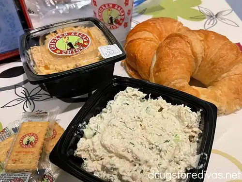 Two small tubs of chicken salad, crackers, croissants, and a cup with water on a table in a Chicken Salad Chick restaurant.