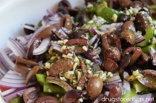 Dressing on top of cucumber, olives, red onion, green pepper, and tomato pieces in a white bowl.