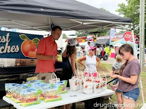 A woman in a purple shirt and shorts looking at a business card at a vendor stand that's selling peaches at the NC Peach Festival in Candor, NC.