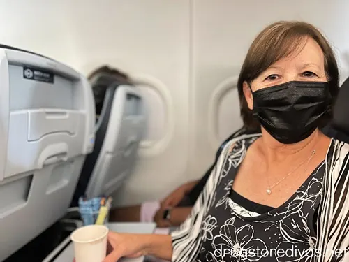 A woman wearing a mask sitting on an airplane and holding a coffee cup.