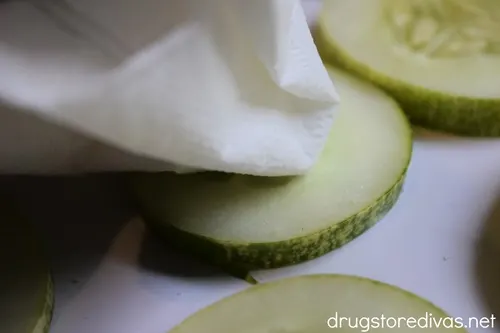 A napkin blotting water off cucumber slices on a white tray.