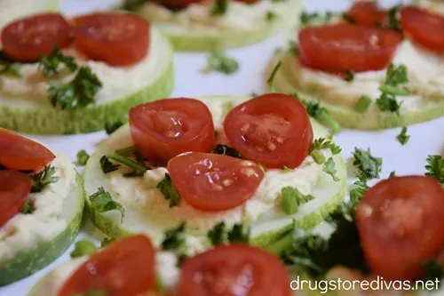 Cherry tomato slices, chopped parsley, and hummus on top of cucumber slices on a white serving tray.