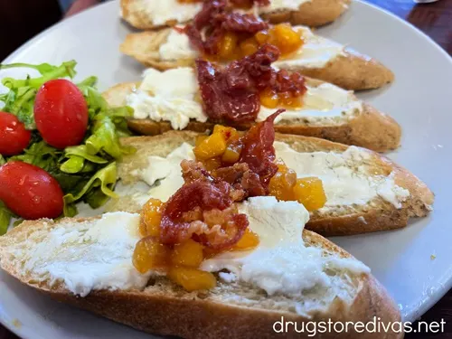 Five crustinis with goat cheese, peach, and prosciutto on a plate with green lettuce and tomatoes.