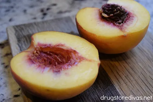 A peach, cut in half, on a wooden cutting board.