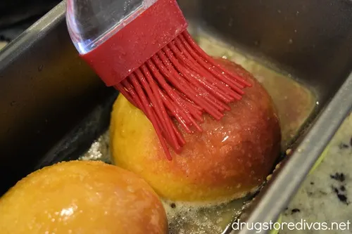 Two peach halves in a bread pan with a pastry brush brushing butter on top.