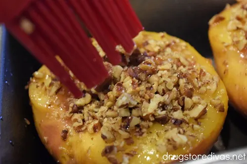 A peach half with chopped pecans on top getting brushed with a pastry brush.