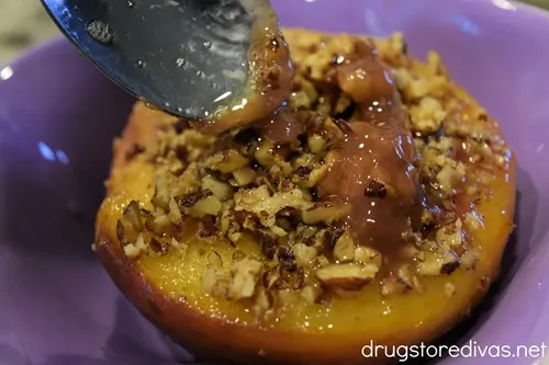 A spoon spooning toffee onto a peach half with chopped pecans on top in a purple bowl.