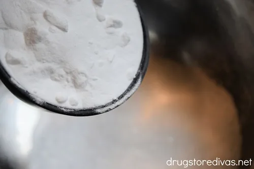 Baking powder in a measuring cup being poured into a silver bowl.