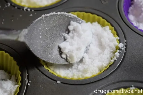 A spoon scooping white powder into a yellow silicone liner in a cupcake mold.