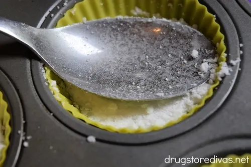 The back of a spoon patting down white powder into a yellow silicone liner in a muffin tin.