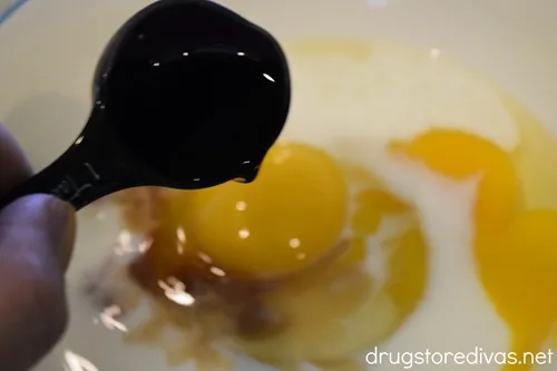A measuring spoon with a dark liquid being poured into a bowl of cracked eggs.
