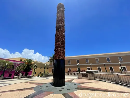 A large cylindrical statue with a sundial looking painting below it surrounded by buildings in Plaza del Quinto Centenario in San Juan, Puerto Rico.