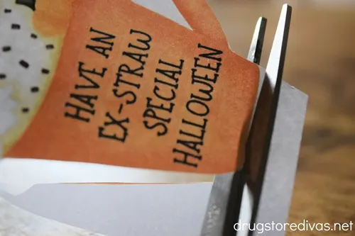 Scissors cutting a cup that says "Have An Ex-Straw Special Halloween" out of a piece of paper.