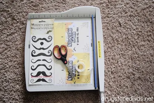 A paper trimmer, a strip of mustache tattoos, scissors, tape, and Halloween cards on a carpet.