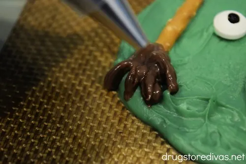 A piping tip piping a broom's bristles on the end of a pretzel stick that's on melted green chocolate.