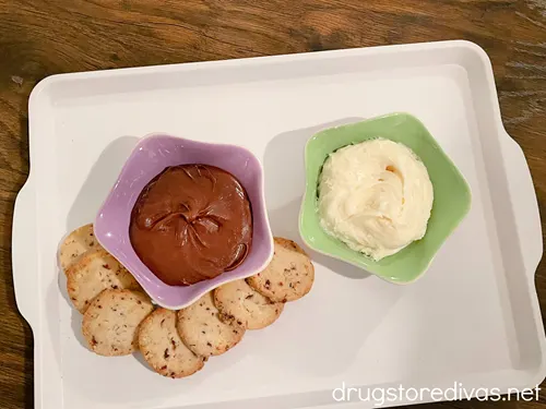 A white tray with chocolate icing in a purple bowl, white icing in a green bowl, and a few cookies.