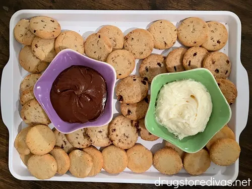 A white tray with chocolate icing in a purple bowl, white icing in a green bowl, and filled with cookies.