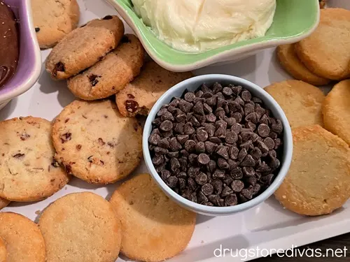 A green bowl with white icing and a blue bowl with mini chocolate chips surrounded by cookies on a white tray.