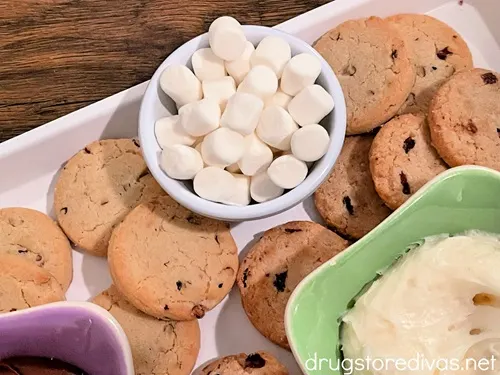 A green bowl with white icing and a blue bowl with mini marshmallows surrounded by cookies on a white tray.