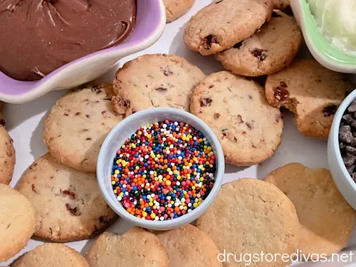 A purple bowl with chocolate icing and a blue bowl with sprinkles surrounded by cookies on a white tray.