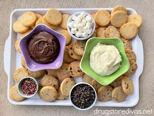 A cookie charcuterie board: A white tray filled with cookies and bowls with chocolate icing, white icing, mini marshmallows, mini chocolate chips, and sprinkles.