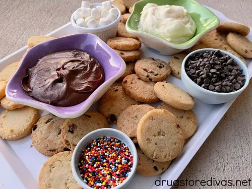 A white tray filled with cookies and bowls with chocolate icing, white icing, mini marshmallows, mini chocolate chips, and sprinkles.