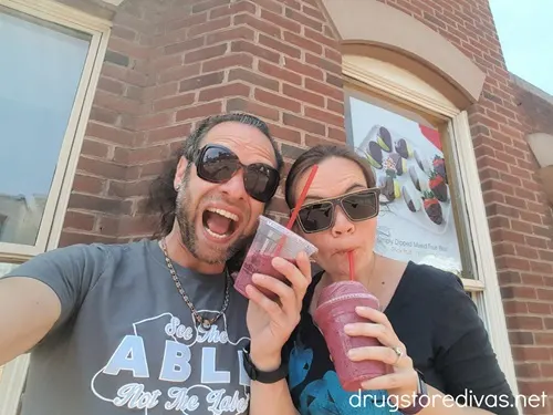 A man and a woman drinking smoothies in front of a brick wall of the store Edible Arrangements.
