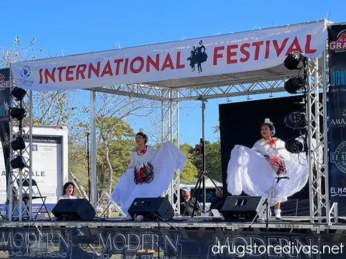 Two female dancers in white dresses on a stage with a banner that says "International Festival" above them.