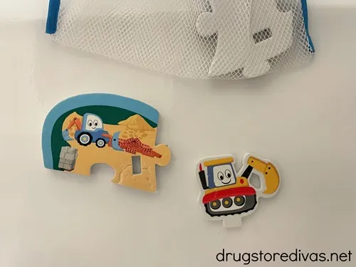 Two foam bath stickers - one is a cartoon bulldozer and one is a cartoon construction vehicle that's on a puzzle piece - on the side of a tub with a mesh bag with a bath sticker on top.