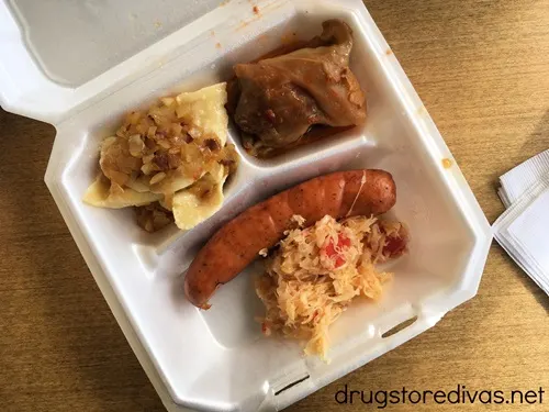 A Styrofoam container with pierogies with onions, a cabbage roll, kielbasi, and sauerkraut on it.