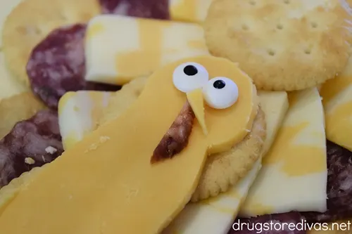 A cheese slice that looks like a turkey face with two candy eyes, a nose, and wattle on top of cheese and crackers.