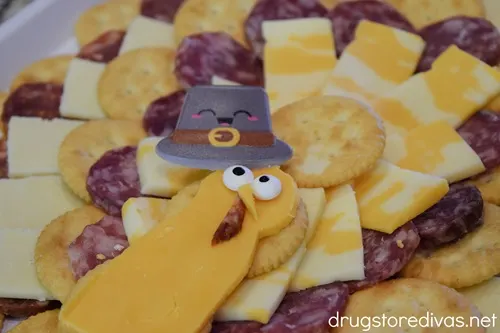 A cheese slice that looks like a turkey face with two candy eyes, a nose, and wattle, with a plastic pilgrim hat, on top of cheese and crackers.