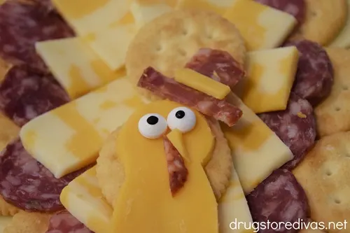 A cheese slice that looks like a turkey face with two candy eyes, a nose, and wattle, with a cheese and salami pilgrim hat, on top of cheese and crackers.