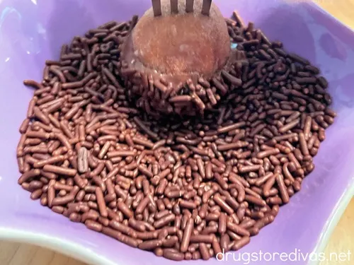A donut hole on a stick being dipped into chocolate sprinkles in a purple bowl.