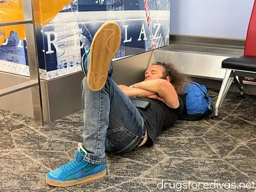 A man in a black shirt, jeans, and teal sneakers taking a nap on an airport floor while using his carry on as a pillow.