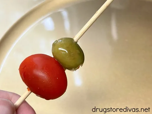 A grape tomato and a green olive on a skewer over a gold plate.