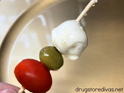 A grape tomato, a green olive, and a mozzarella ball on a skewer over a gold plate.