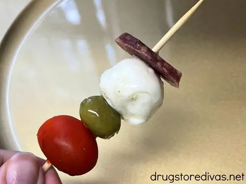 A grape tomato, a green olive, a mozzarella ball, and a slice of salami on a skewer over a gold plate.