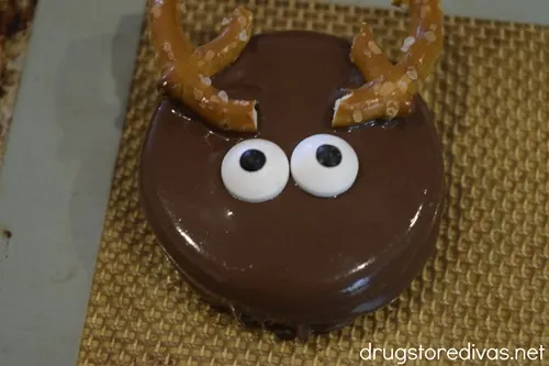 A dipped Oreo cookie with pretzel antlers and two candy eyes on a silicone baking mat.
