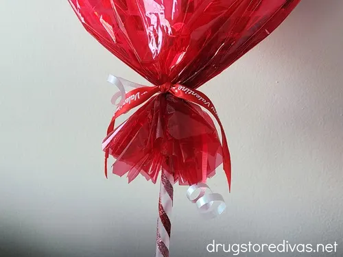 A heart-shaped lollipop in red cellophane that's gathered near the stick and tied with white curling ribbon and red ribbon.
