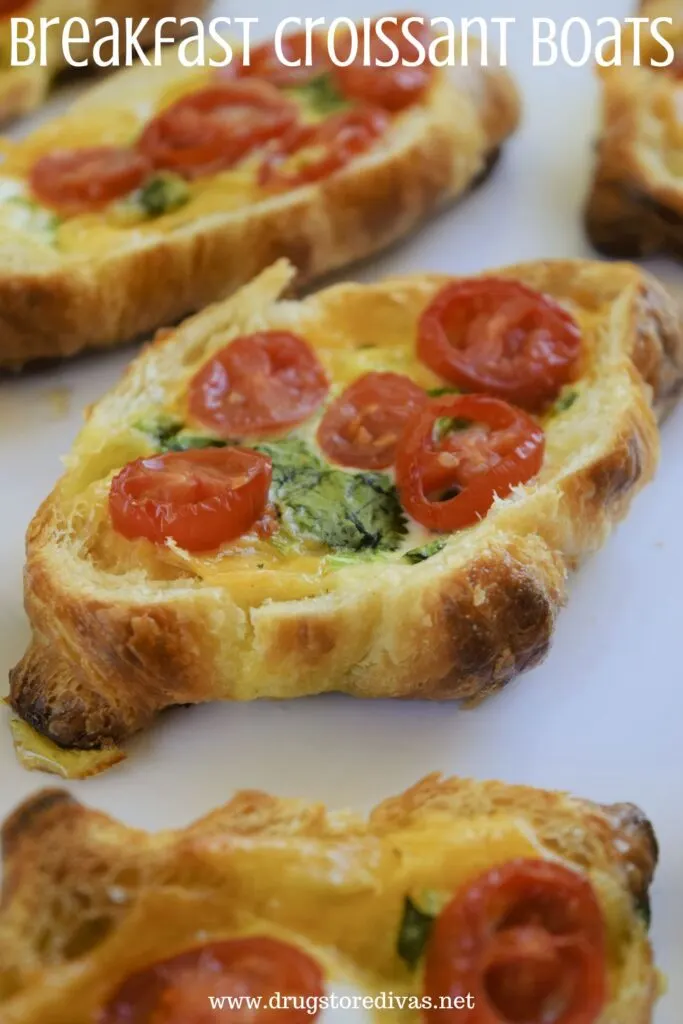 Five mini croissants with the tops cut off, filled with eggs, cilantro, and grape tomato slices on a white tray with the words "Breakfast Croissant Boats" digitally written on top.