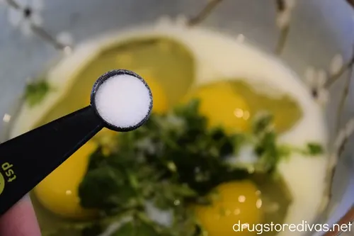 A measuring spoon above a blue bowl with milk, four cracked eggs, and chopped cilantro.