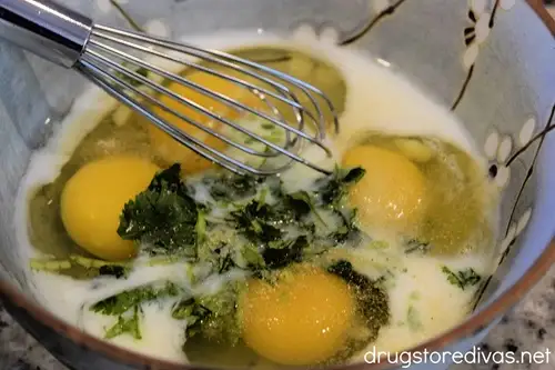 A whisk in a blue bowl with milk, four cracked eggs, and chopped cilantro.