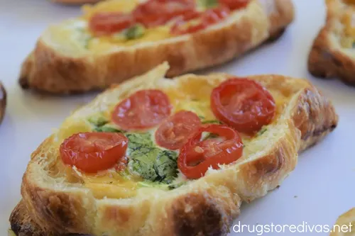 Four baked Breakfast Croissant Boats, which are croissants cut open with cooked eggs, herbs, and sliced grape tomatoes on top, on a white tray.