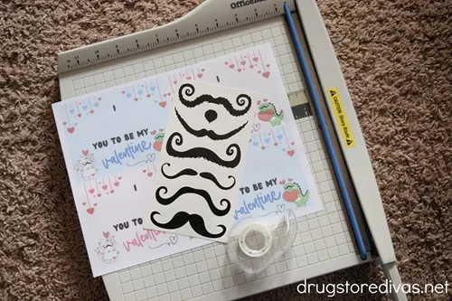 A printable of Valentine's Day cards, mustache tattoos, and tape on top of a paper trimmer.