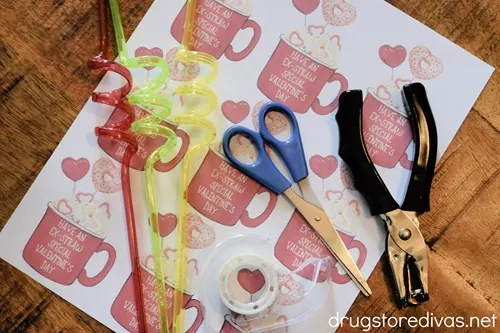 A printable Valentine's Day tag sheet, three crazy straws, scissors, tape, and a hole punch on a table.